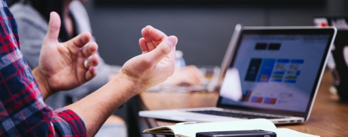 A imagem apresenta do lado esquerdo, um profissional argumentando com as mãos com seu colega de trabalho as informações apresentadas na tela do notebook.